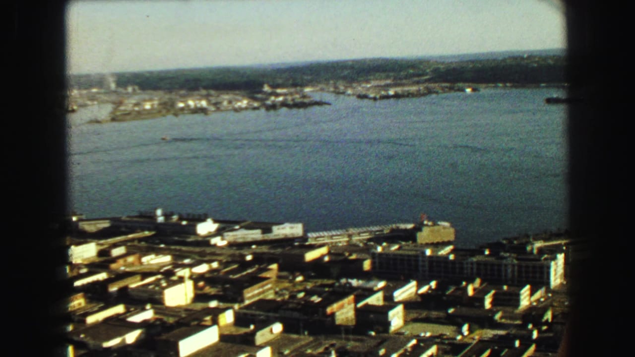 1968 Vancouver harbor frame showing open water, dockside buildings, a distant shoreline, and a small smoke or steam plume.
