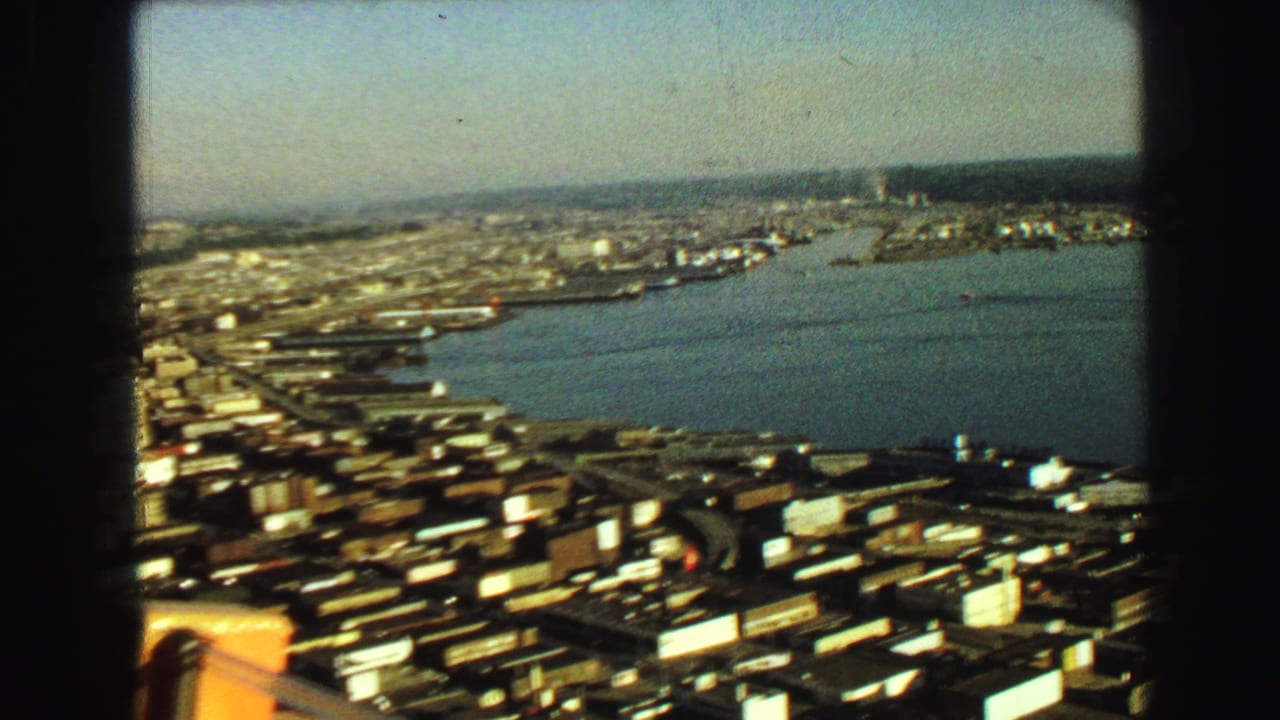 Aerial view over a waterfront city with a wide waterway and industrial shoreline.