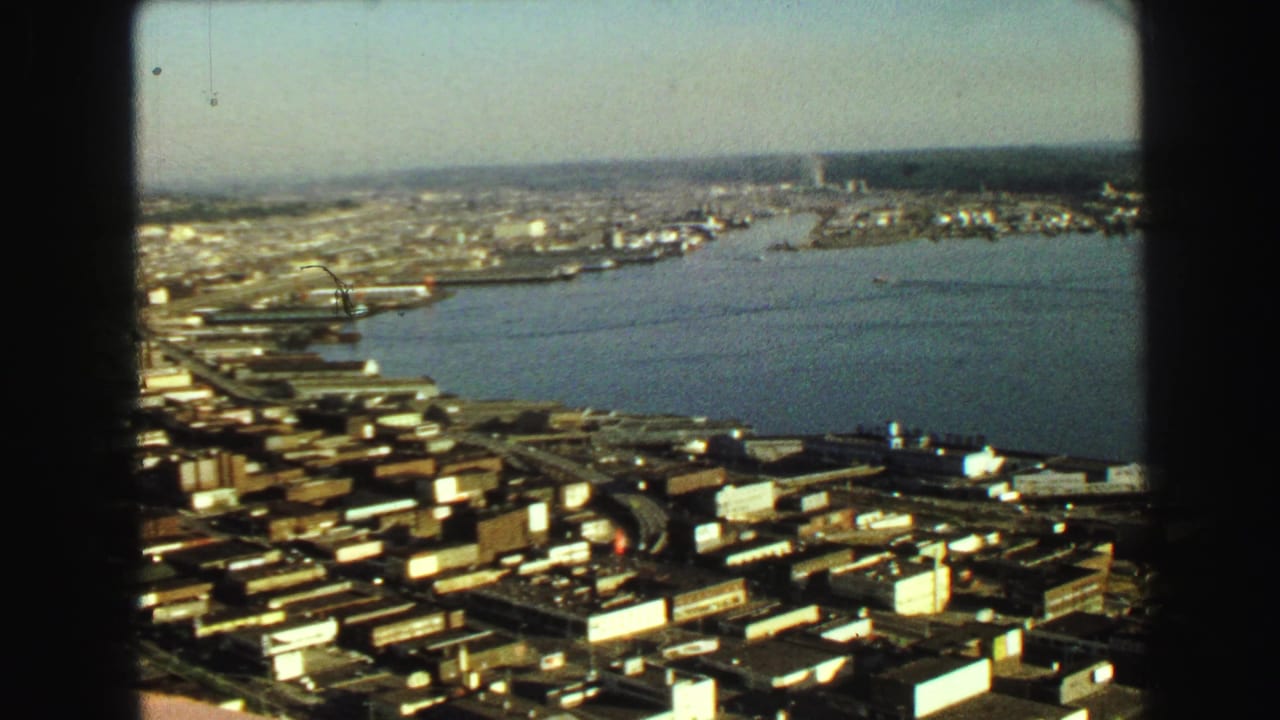 Aerial city view with harbor water, docks, low buildings, and dark film frame edges.