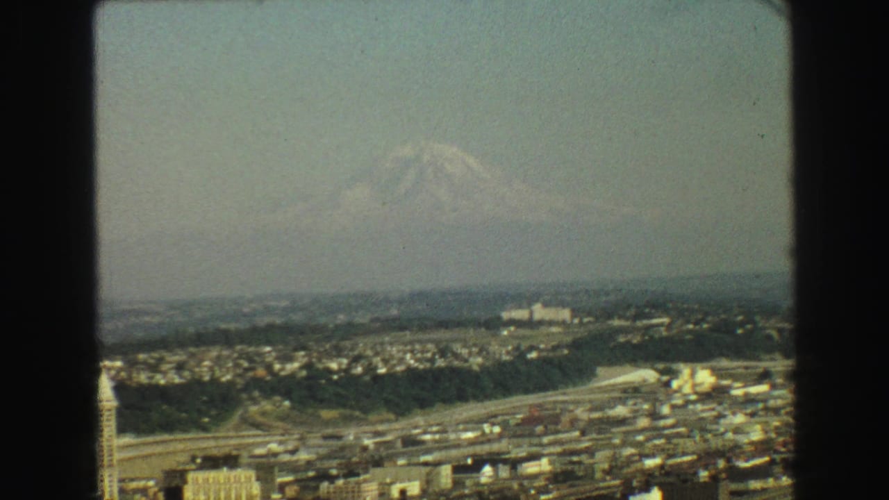 Hazy city view with a distant snow-covered mountain beyond roads and low buildings.