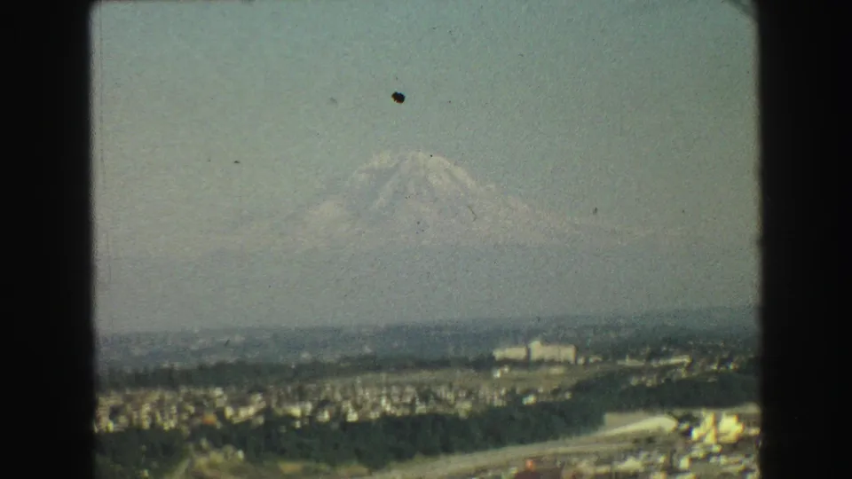 1968 8mm frame of a Vancouver-area skyline view, with a snow-covered mountain rising behind the city haze.