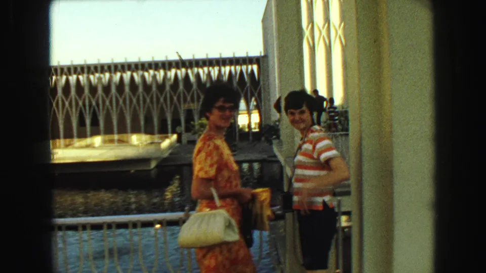 1968 Vancouver frame showing two women posing by a column, with water, railings, and white lattice architecture behind them.