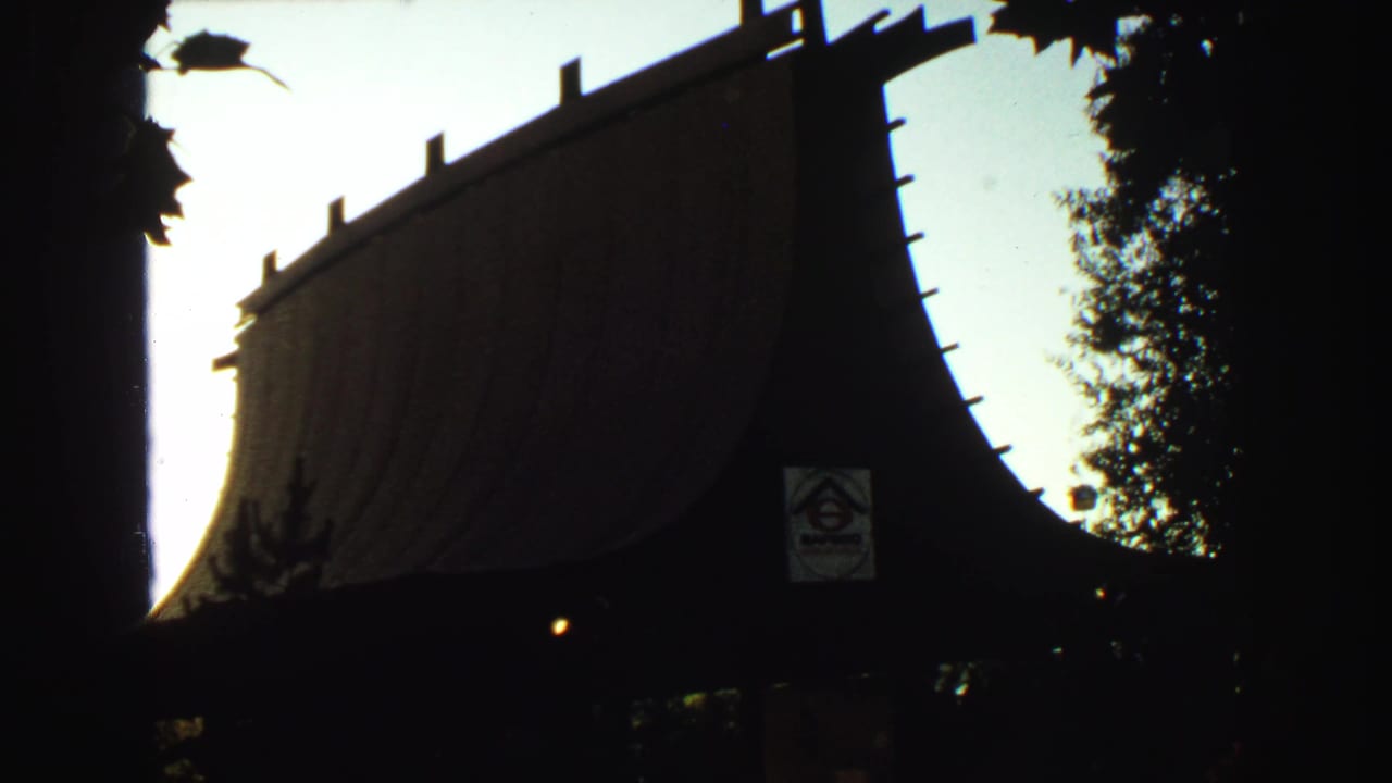 1960s 8mm frame showing a dark Japanese-style roofline or gate near trees, with a small posted sign visible in silhouette.