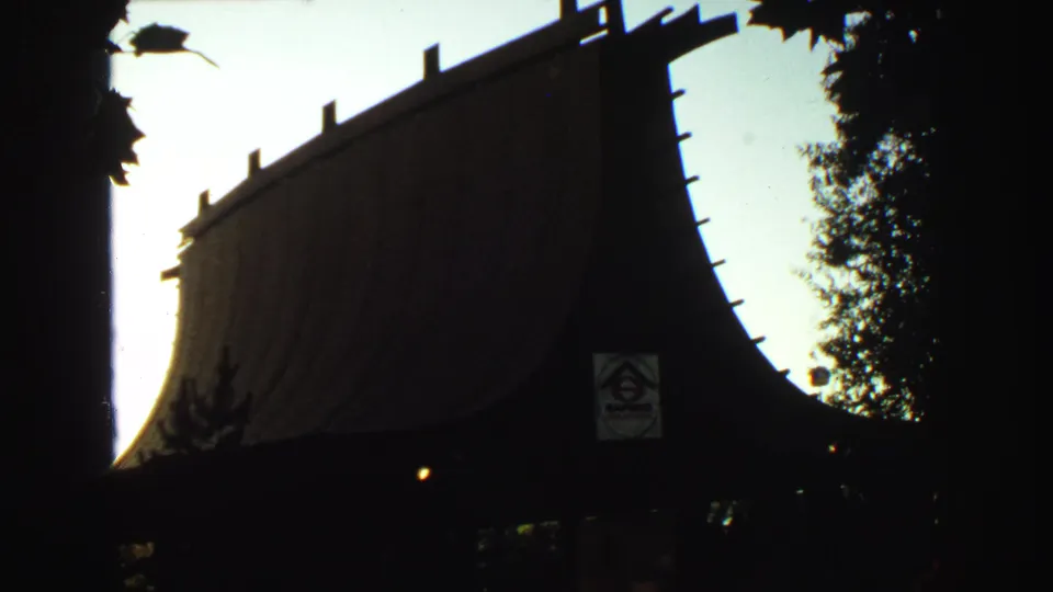 1960s 8mm frame showing a dark Japanese-style roofline or gate near trees, with a small posted sign visible in silhouette.