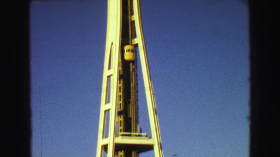 1960s 8mm low-angle view of the Space Needle structure with a yellow elevator car rising against a clear blue sky.