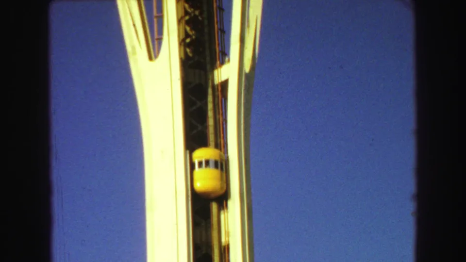 1968 8mm frame of a yellow elevator capsule on a tall observation-tower shaft, framed by white supports against clear blue sky.