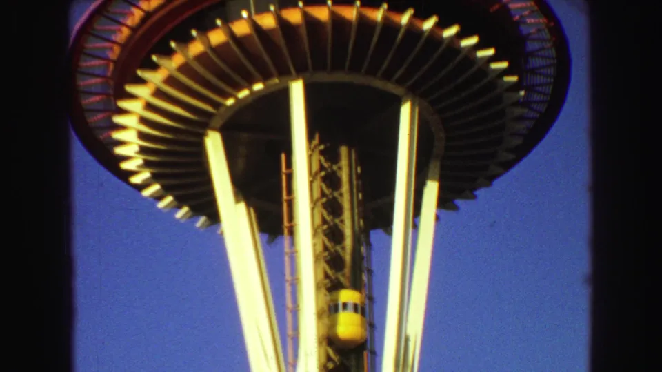 8mm frame of the yellow elevator beneath a circular observation deck, with white tower legs and blue sky in the background.