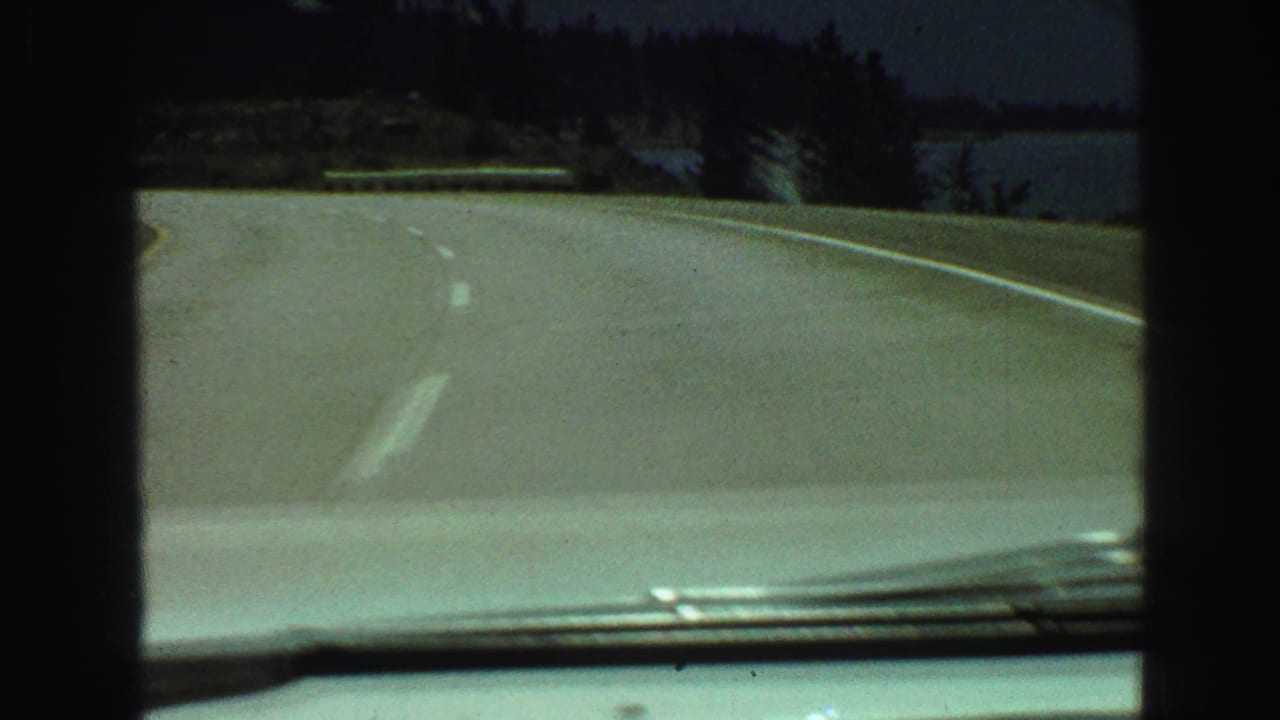 View over a car hood on a curving paved road bordered by trees and water.