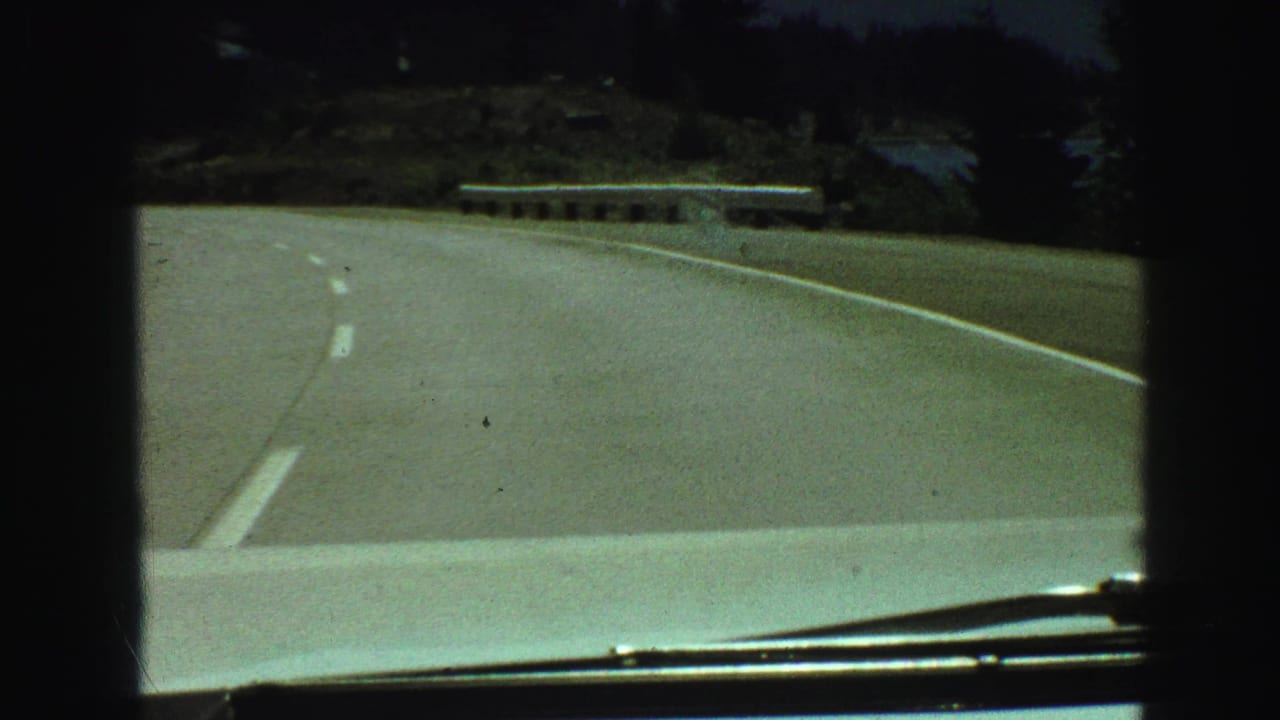 Car approaches a bend in a paved road with trees and water beyond the shoulder.