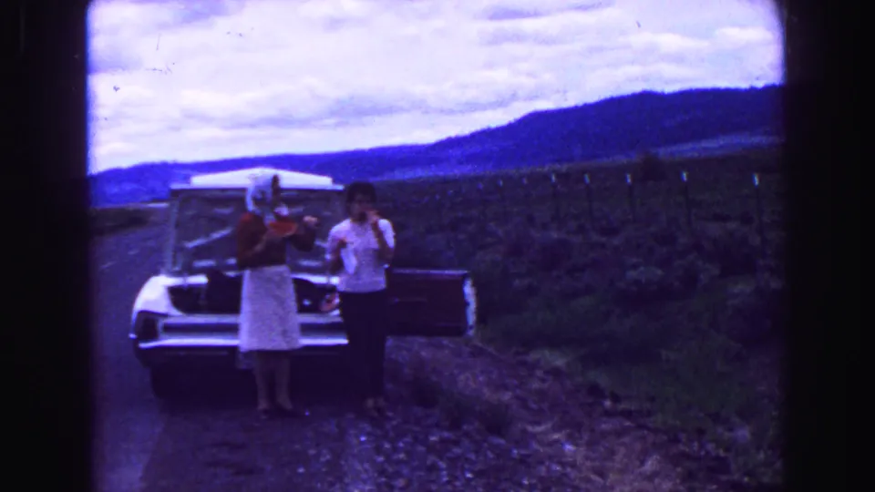 Two adults stand by a light-colored car on a rural road, holding watermelon in 1968 home-movie footage.