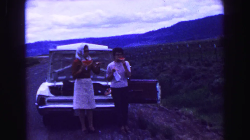 Two adults eat watermelon beside a car on a rural British Columbia roadside, with open fields and hills behind them.