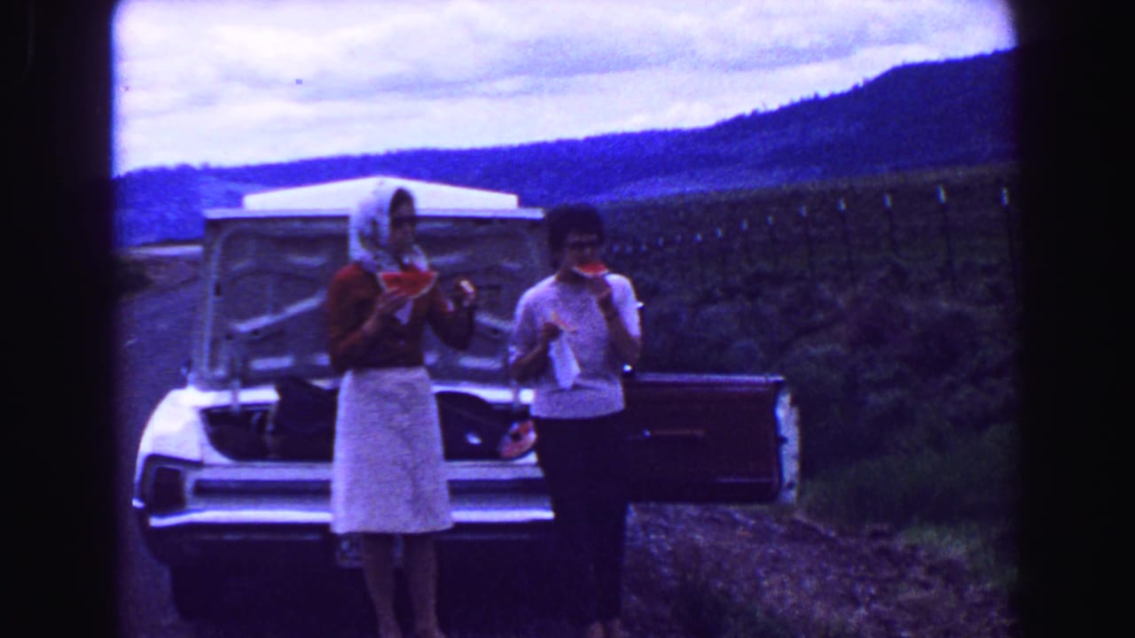1968 8mm home-movie frame of two women eating watermelon near an open car trunk during a mountain road trip.
