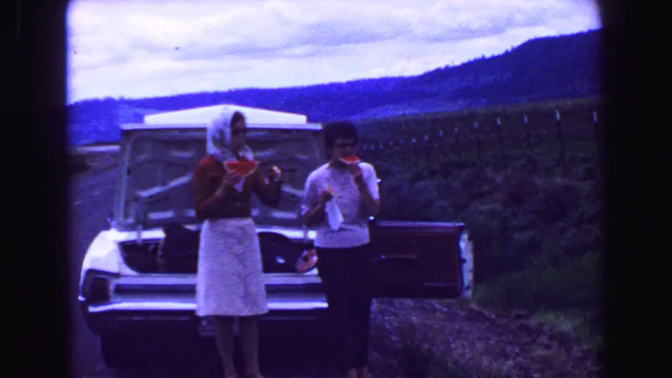 1968 8mm home-movie frame of two women eating watermelon near an open car trunk during a mountain road trip.