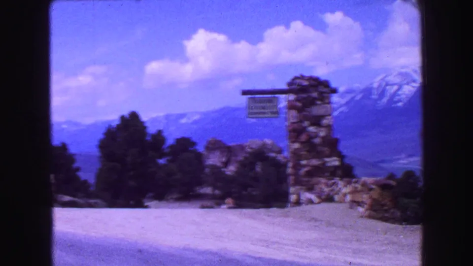 1968 8mm frame showing a Geiger Lookout sign mounted on stonework, with evergreens and snow-capped mountains behind.