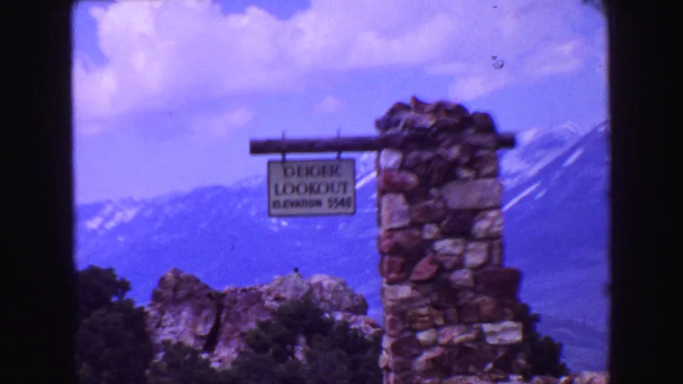 1968 8mm frame of the Geiger Lookout elevation sign on a stone structure, with blue sky and mountains in the background.