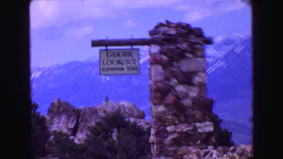 Handheld 8mm frame showing the Geiger Lookout sign and stone marker with rocky foreground and mountain backdrop.