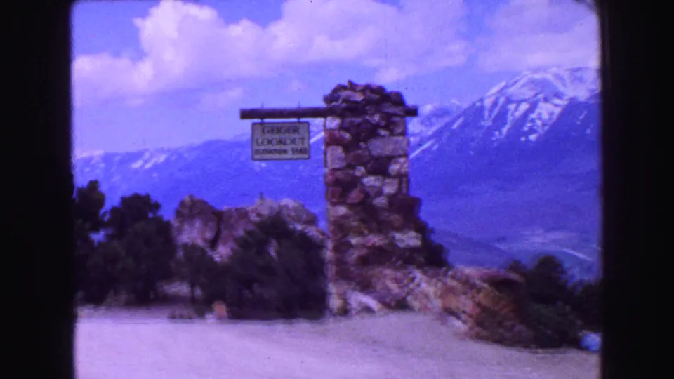 Wider 1968 travel frame of the lookout marker with shrubs, a road or overlook surface, clouds, and snow-capped mountains.