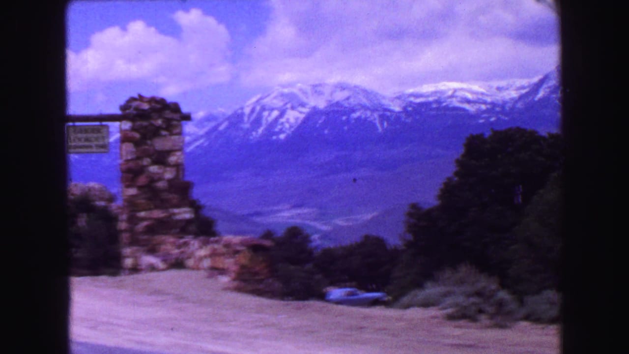 Stone signpost and blue parked car with snow-capped mountains beyond trees.