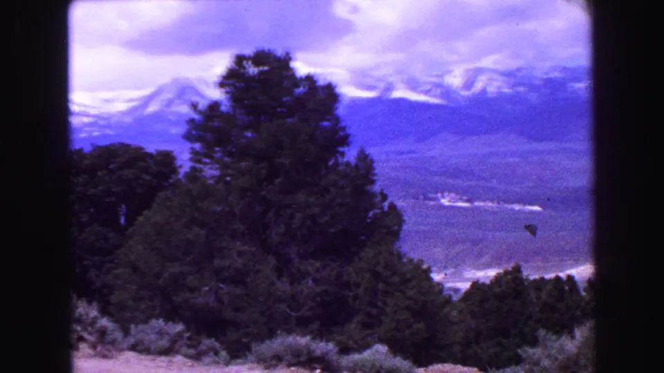 1968 Vancouver-area 8mm frame showing dark evergreens below a blue-purple sky, with distant snow-capped mountains and a broad valley.