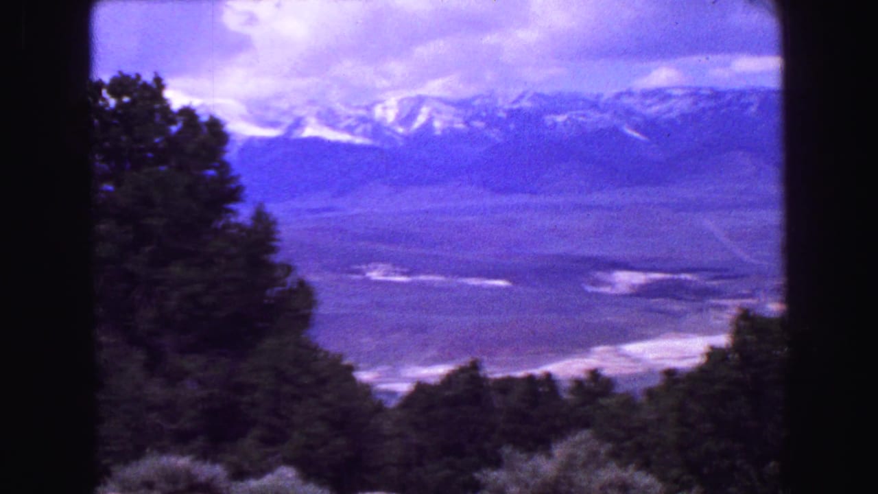 Distant mountain ridge and valley seen past dark foreground trees under a cloudy sky.