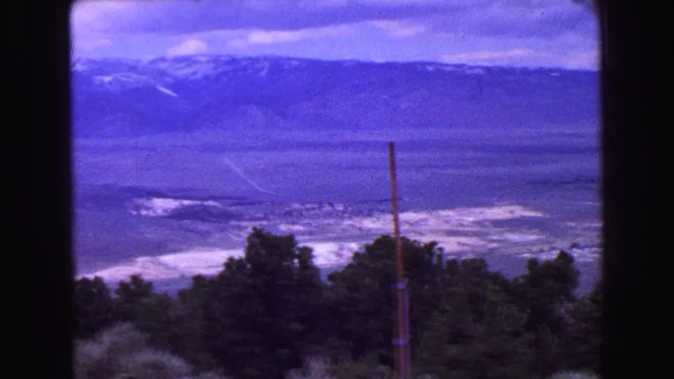 1968 Vancouver-area landscape frame with a distant mountain ridge, valley floor, foreground evergreens, and a slender vertical pole.