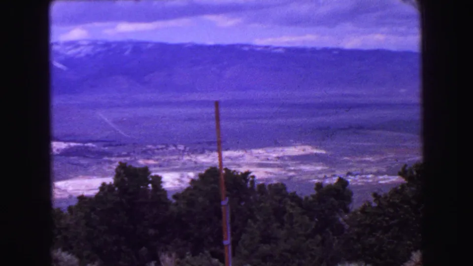1968 8mm frame of a broad valley and distant mountains, with dark trees and a utility pole in the foreground.