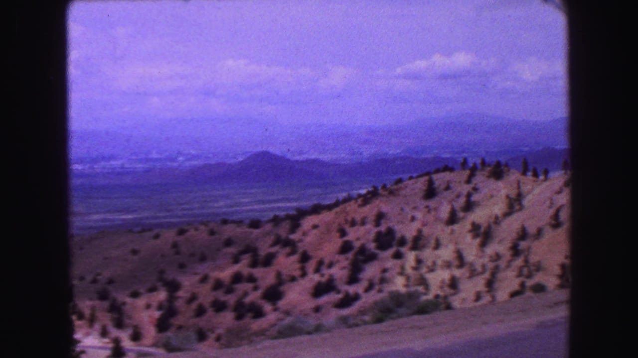 1968 8mm view across a dry, pine-dotted hillside toward hazy mountain ranges under a purple-tinged sky.