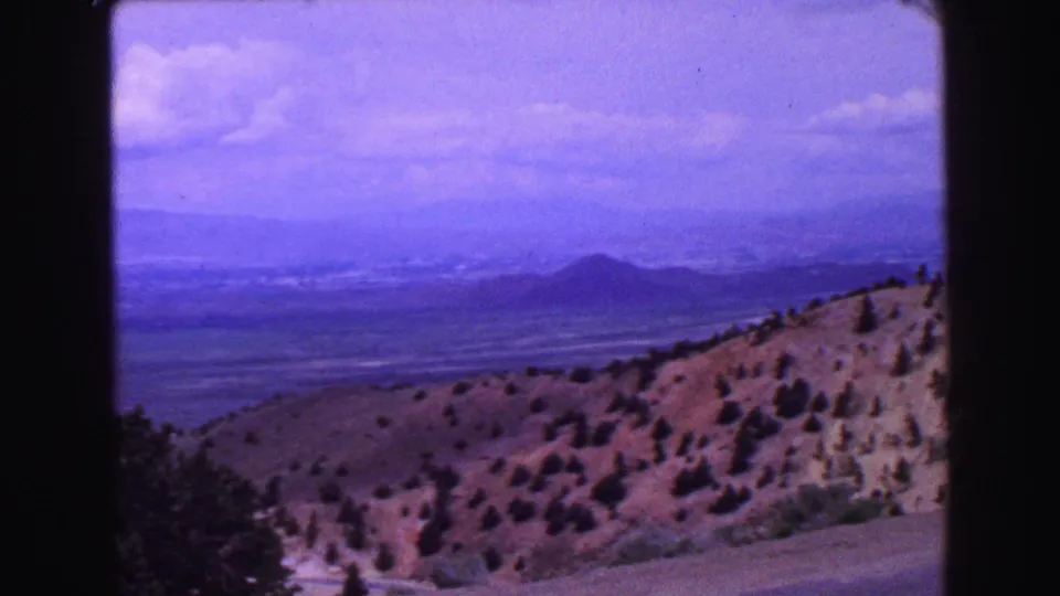 1968 8mm landscape frame showing rolling dry hills, scattered pines, and a hazy mountain horizon.