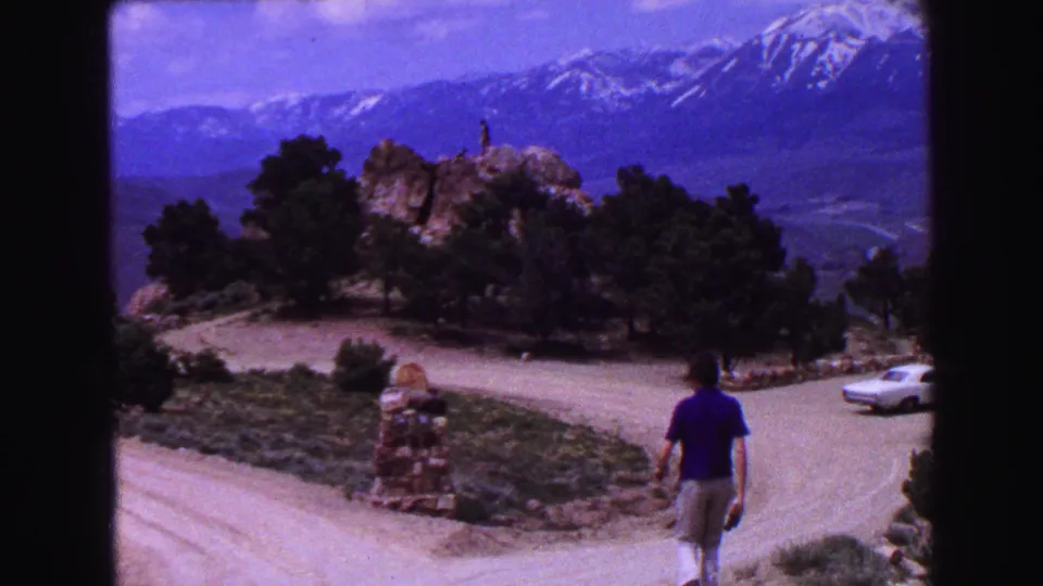 1968 8mm frame of an adult walking by a roadside overlook, with a white car, rocky outcrop, pines, and snowy mountains behind.