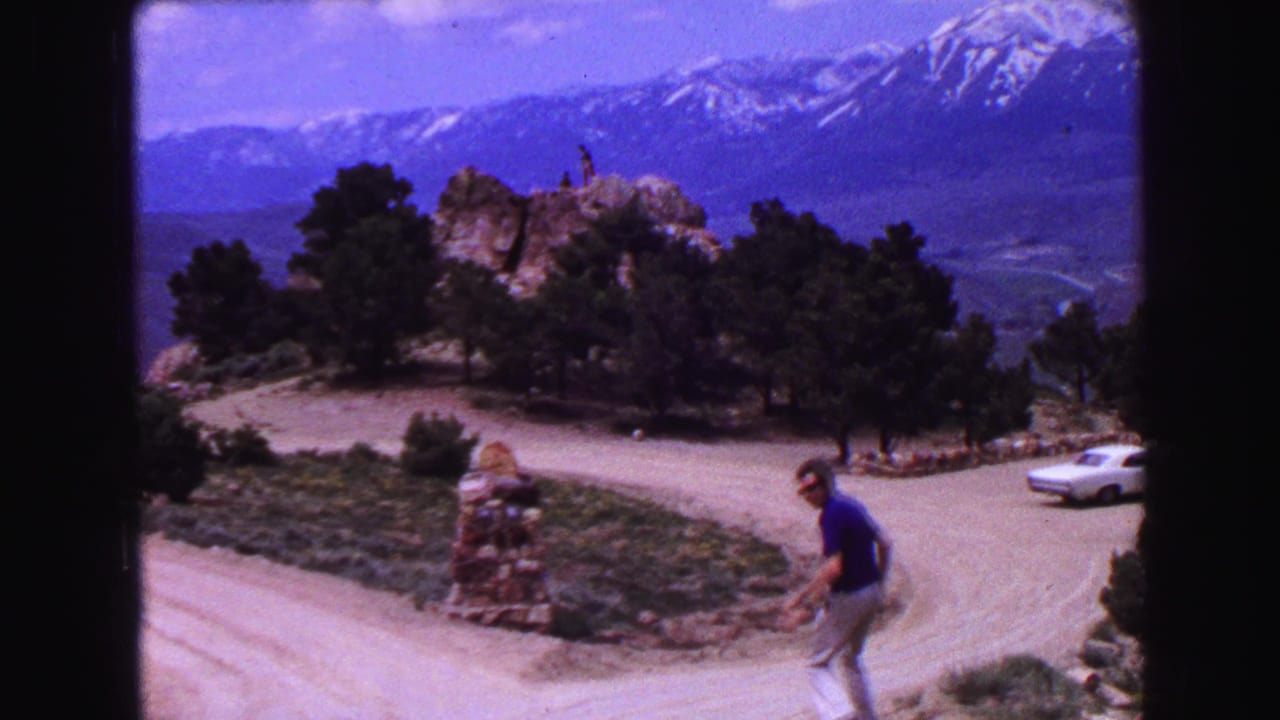 Man walking near a dirt road below a rocky overlook with mountains in the distance.