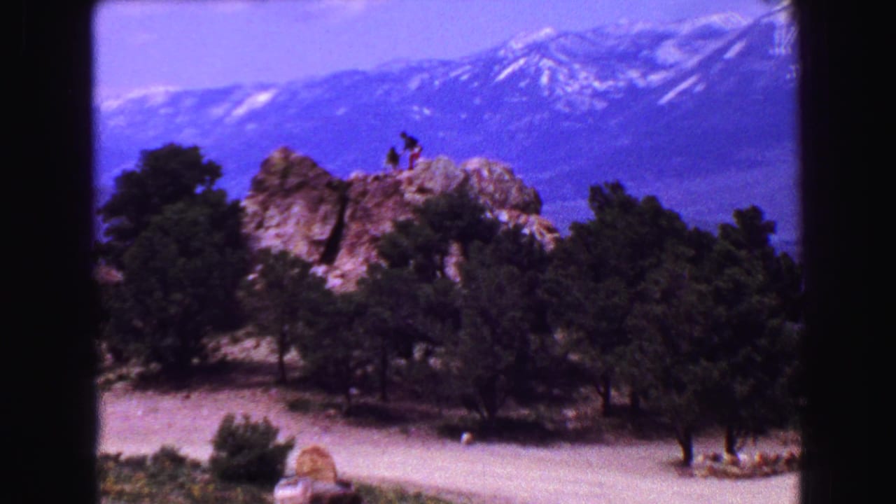 Two people stand on a rocky ledge above trees with snow-capped mountains behind them.