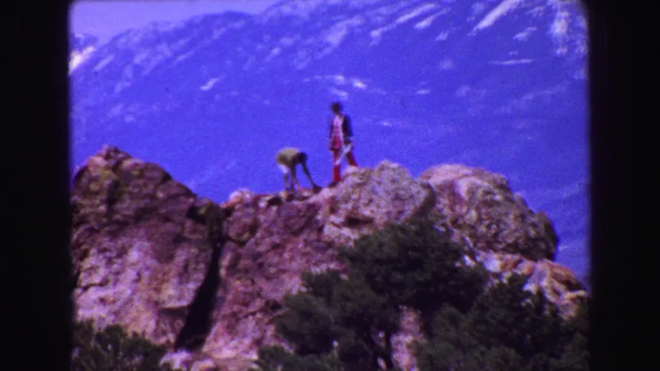 Two visitors stand and bend on a rocky ledge during a 1968 Vancouver-area scenic stop, with purple-blue 8mm color and mountains beyond.