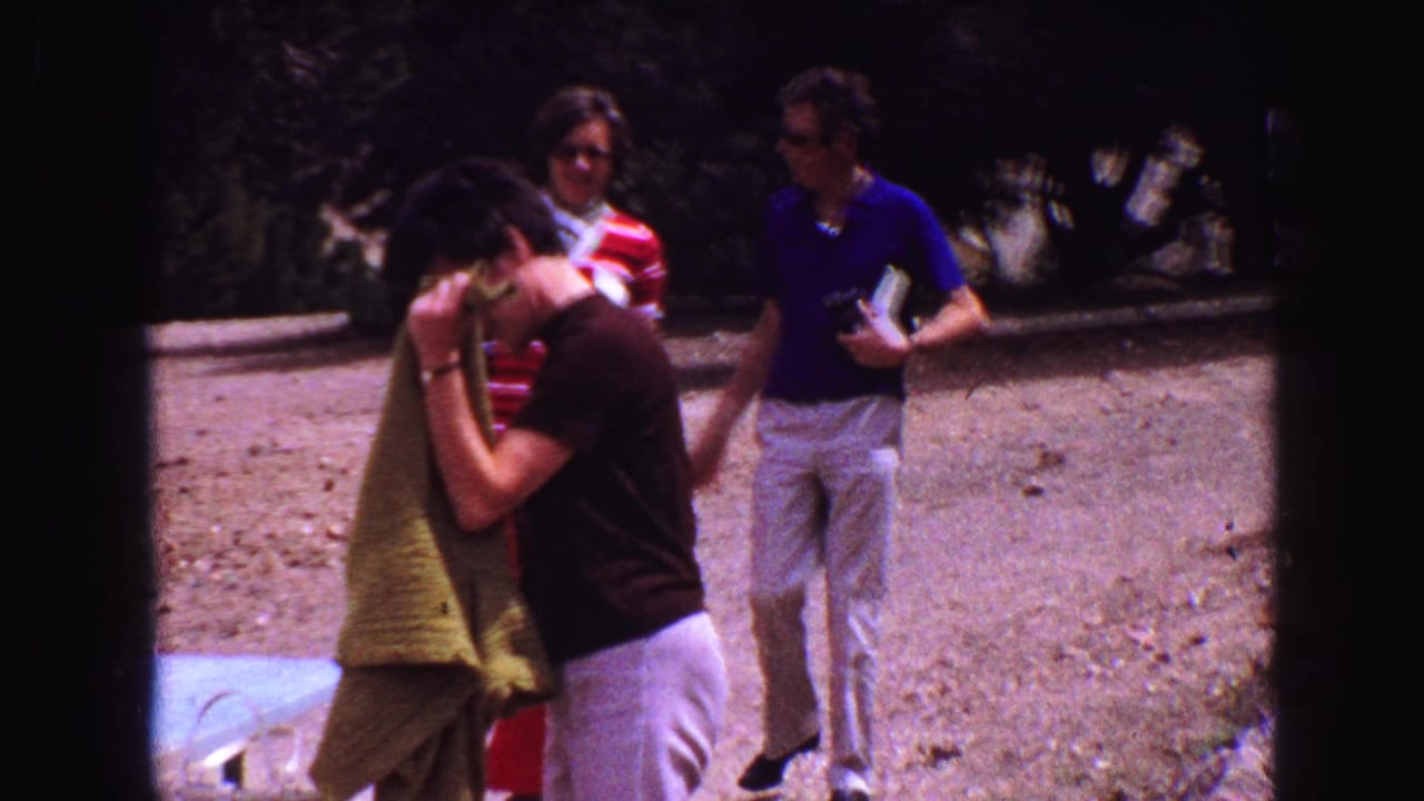 Man folds a green towel near a picnic table as two adults walk behind him.