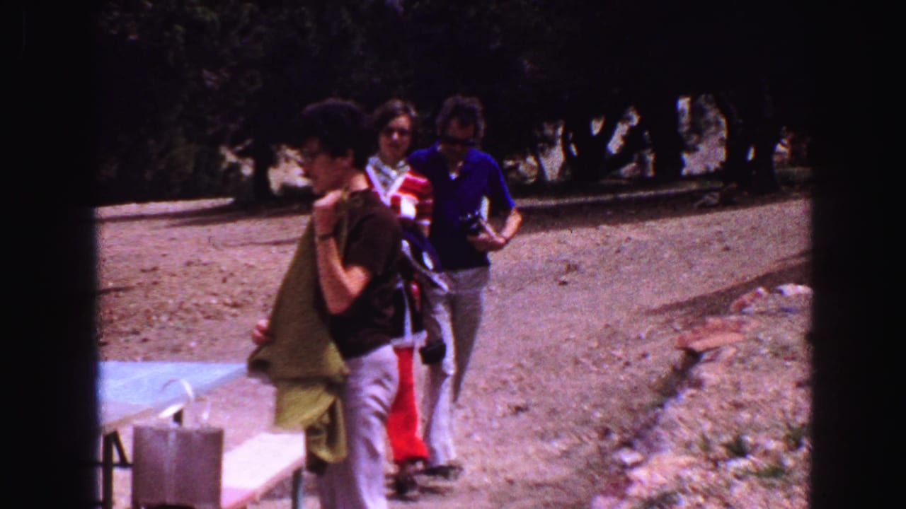 Man holds a towel beside a picnic table while a couple approaches on the path.