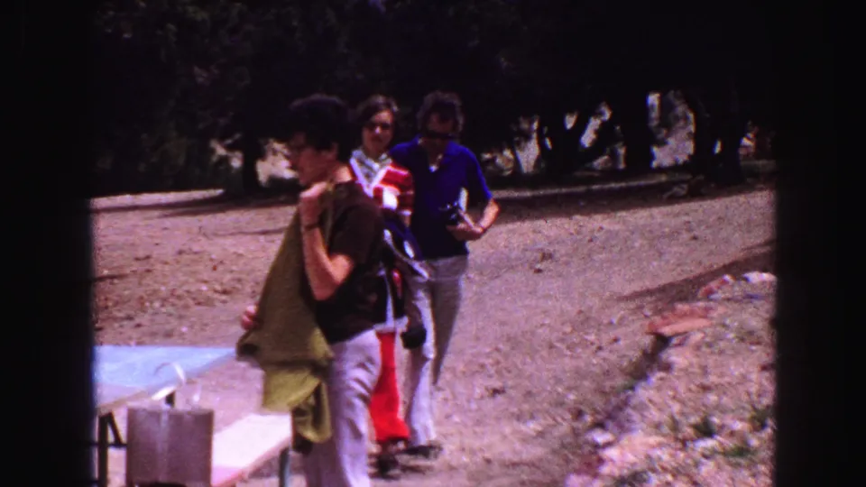 A Vancouver 1968 park scene shows a man with a green towel near picnic benches as two adults walk behind him in bright sun.