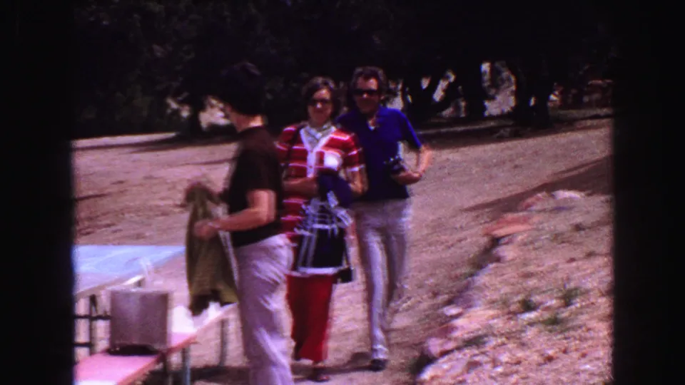 The 1968 Vancouver home-movie frame shows a man turning with a towel beside a picnic table as two adults pass behind on a dirt path.