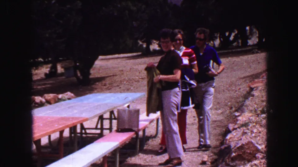 A late-1960s Vancouver park moment shows a man holding a green towel near picnic benches while a couple walks along the path behind him.