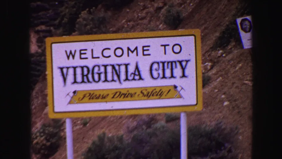 1968 8mm frame showing a yellow and white Welcome to Virginia City sign with Please Drive Safely lettering near a dirt hillside.