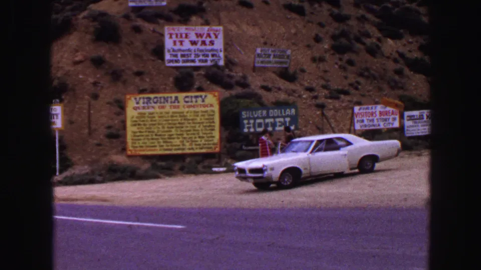 1968 8mm frame showing multiple roadside signs near Virginia City, including Silver Dollar Hotel lettering and a white vintage car by the road.