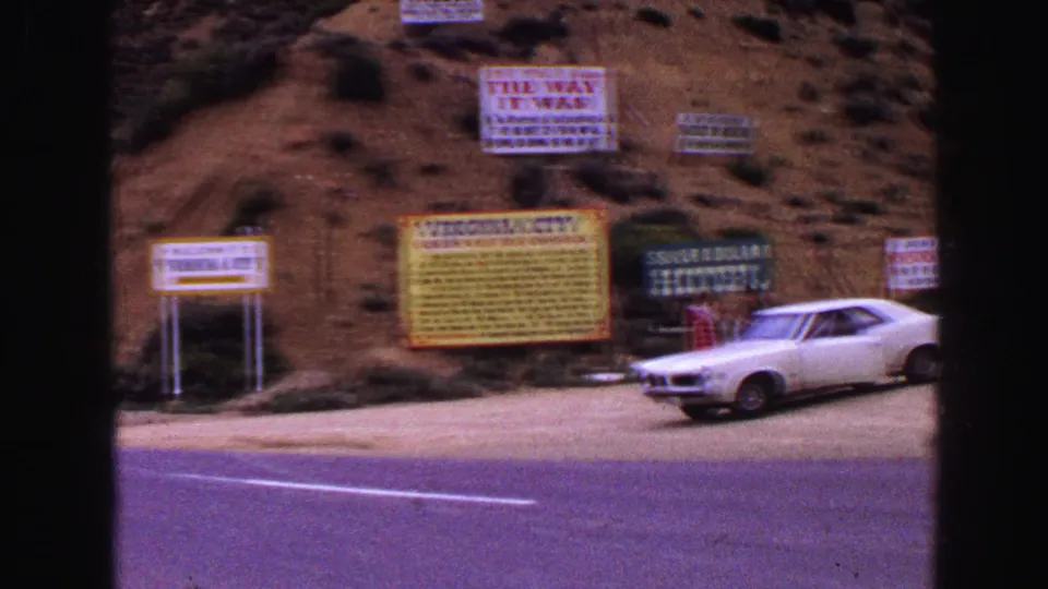 1968 8mm frame with a soft wide view of Virginia City roadside signage, the Silver Dollar Hotel sign, and a white car at frame edge.