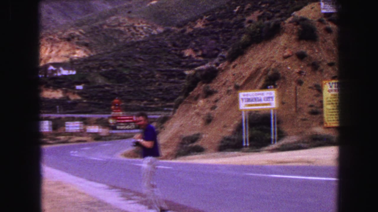 A man in a blue shirt stands near a winding mountain road with Virginia City signage visible in the 1968 Vancouver travel footage.
