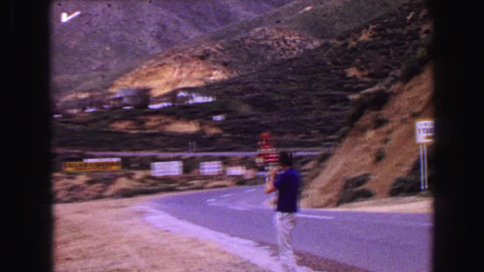 The visitor appears to hold a camera while standing beside the mountain highway, with signs and hillside terrain behind him.