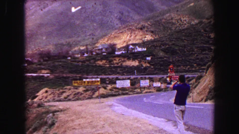 A man photographs the roadside signs from the edge of a winding mountain highway in the 1968 Vancouver home-movie footage.