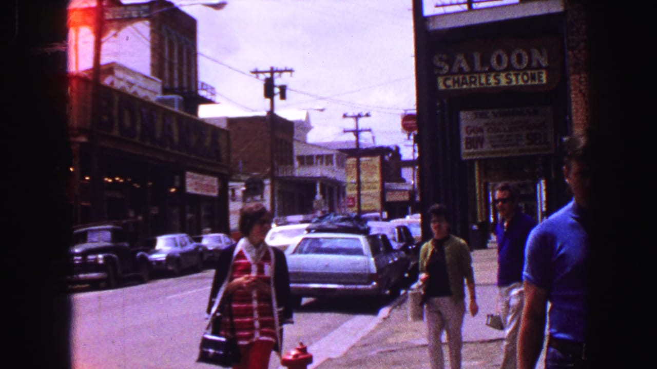 Pedestrians walk along a commercial street with parked cars and storefront signs.