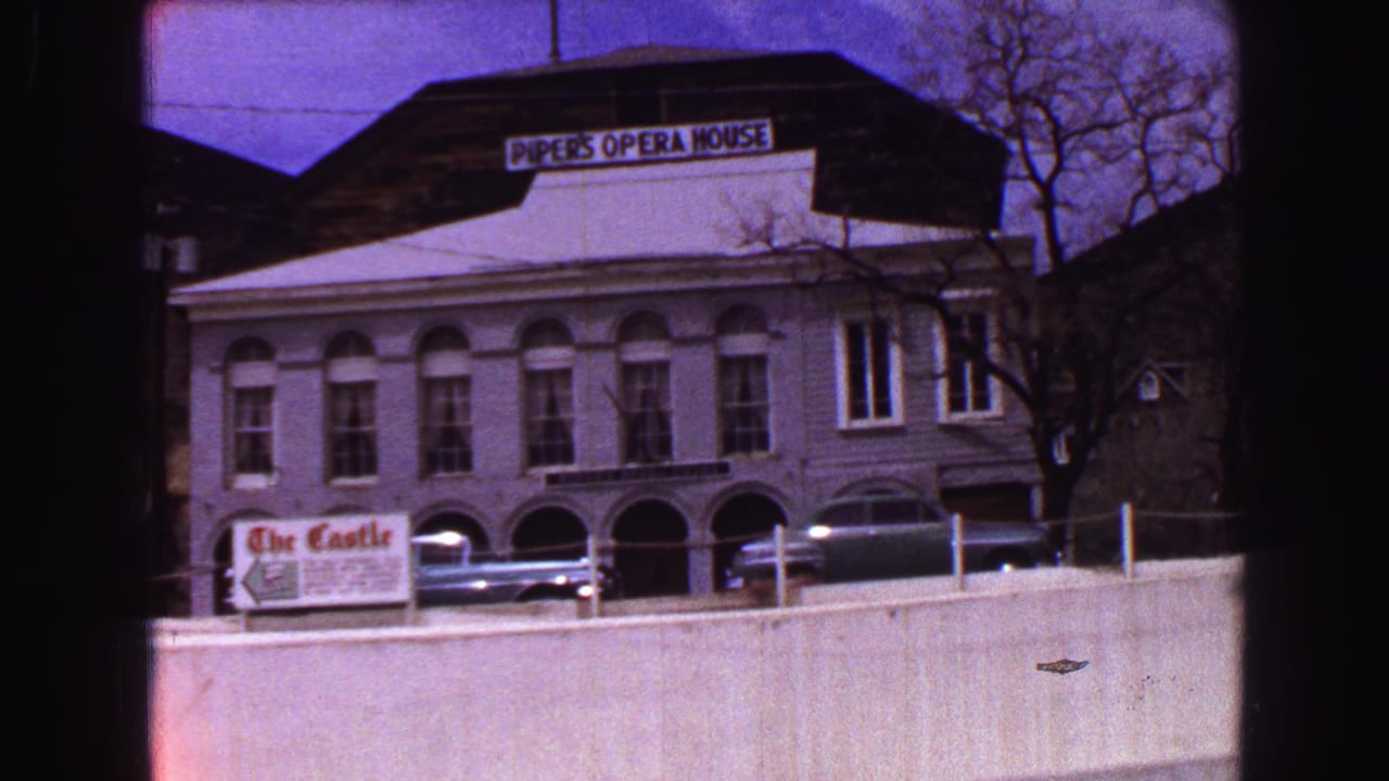 Street-facing view of Piper's Opera House signage, arched windows, parked cars, a low wall, and bare tree branches.