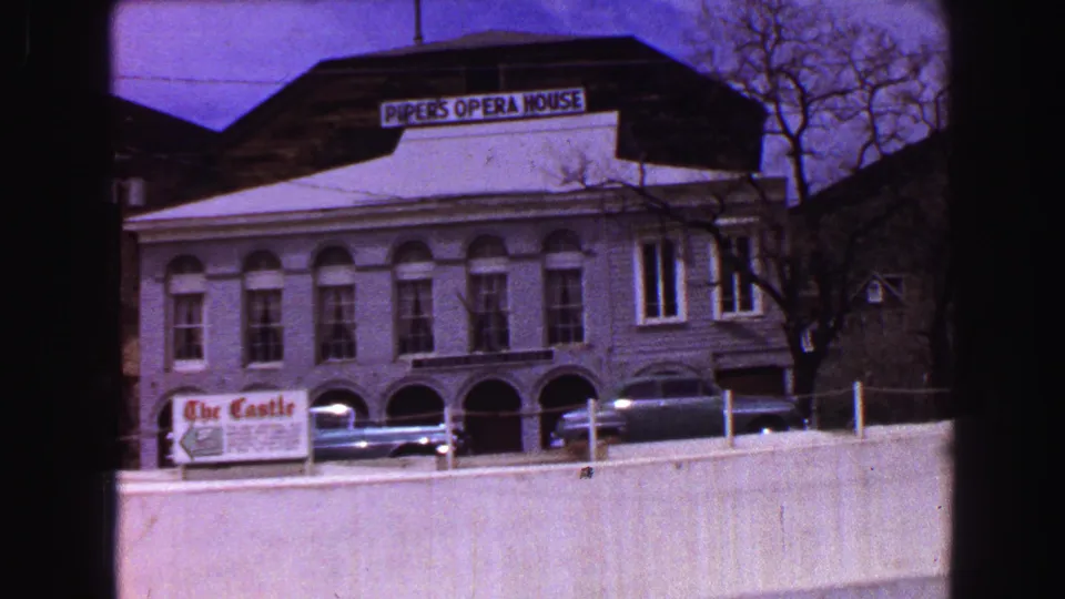 Handheld 8mm view of Piper's Opera House signage, a low wall, arched lower windows, parked cars, and a nearby Castle sign.