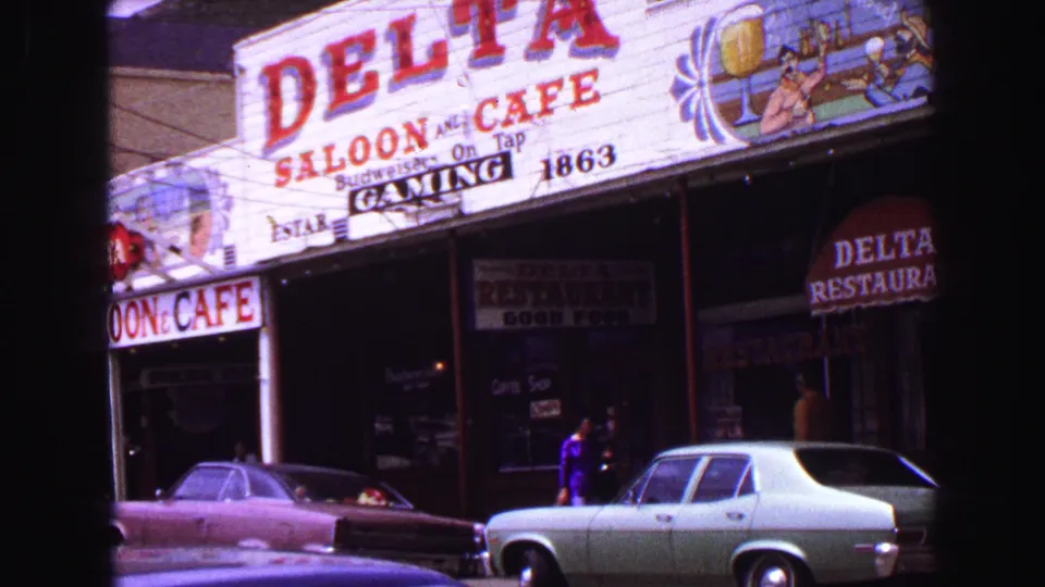 Commercial storefront with Delta Saloon and Cafe signage, parked cars, awnings, and pedestrians on the sidewalk in 1968 Vancouver footage.