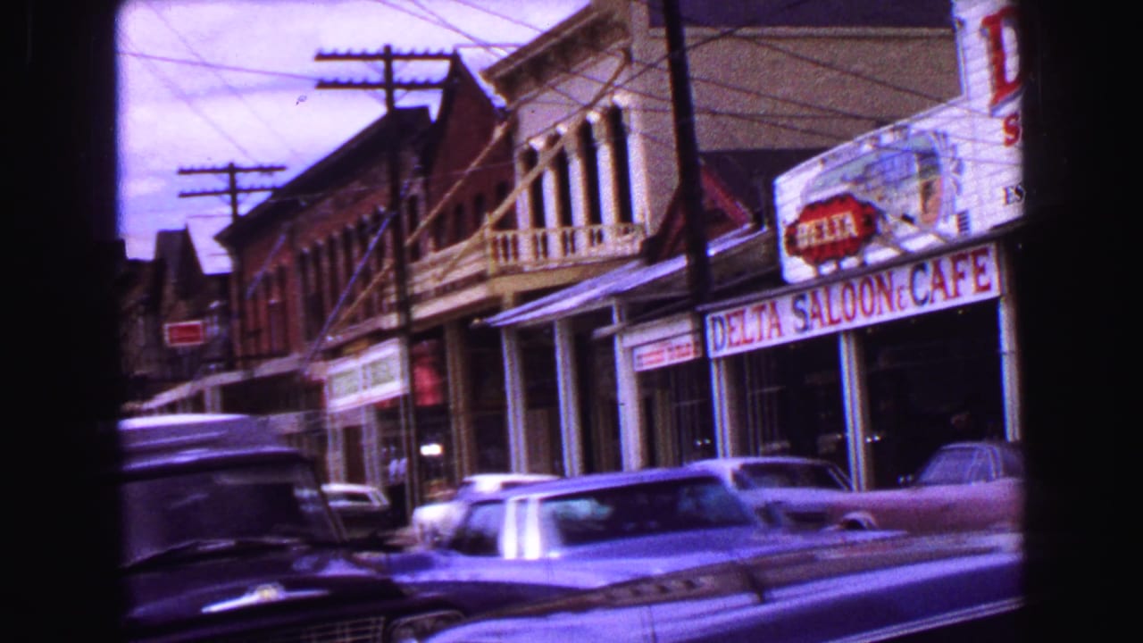 1968 Vancouver 8mm frame showing a tilted street view with storefronts, parked cars, utility poles, and Delta Saloon signage.