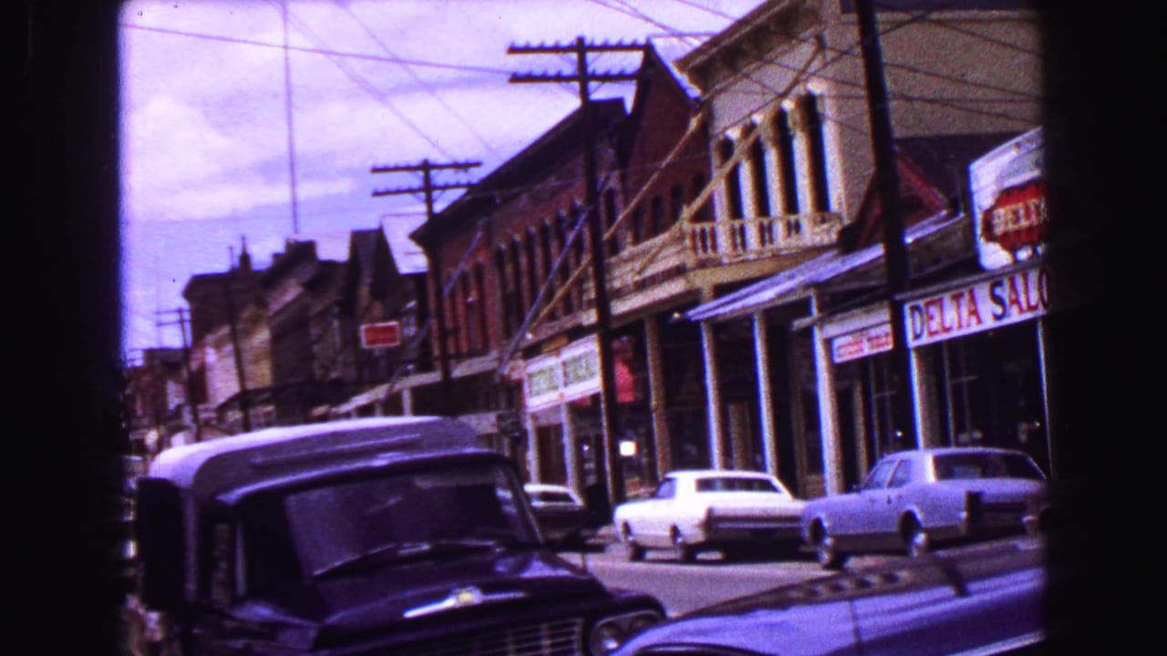 1968 Vancouver-area 8mm frame showing a main street with parked cars, brick buildings, utility poles, and visible saloon frontage.