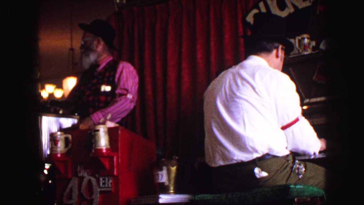 1968 8mm interior frame showing two adult men in top hats near red curtains, bar equipment, glasses, and dim saloon lighting.